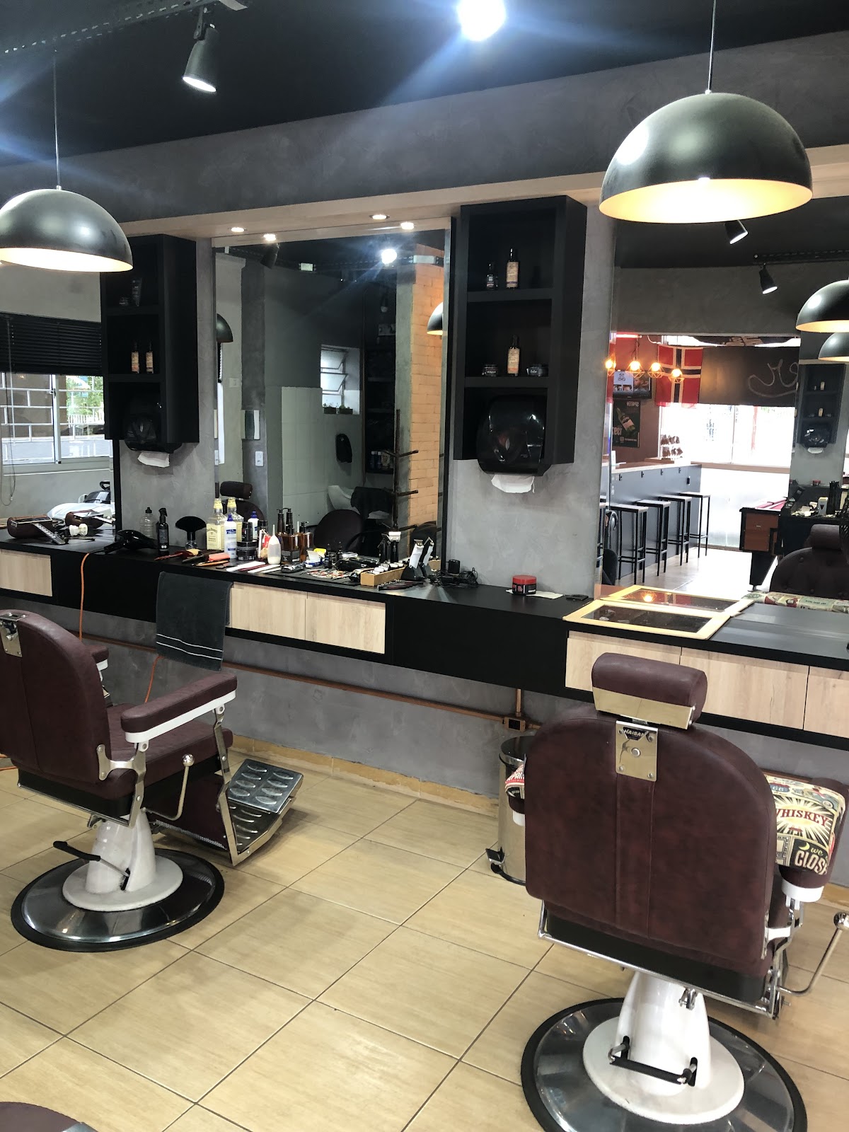 Cadeiras de barbeiro e balcão da Øks Barbearia - Pub, com a área do pub ao fundo