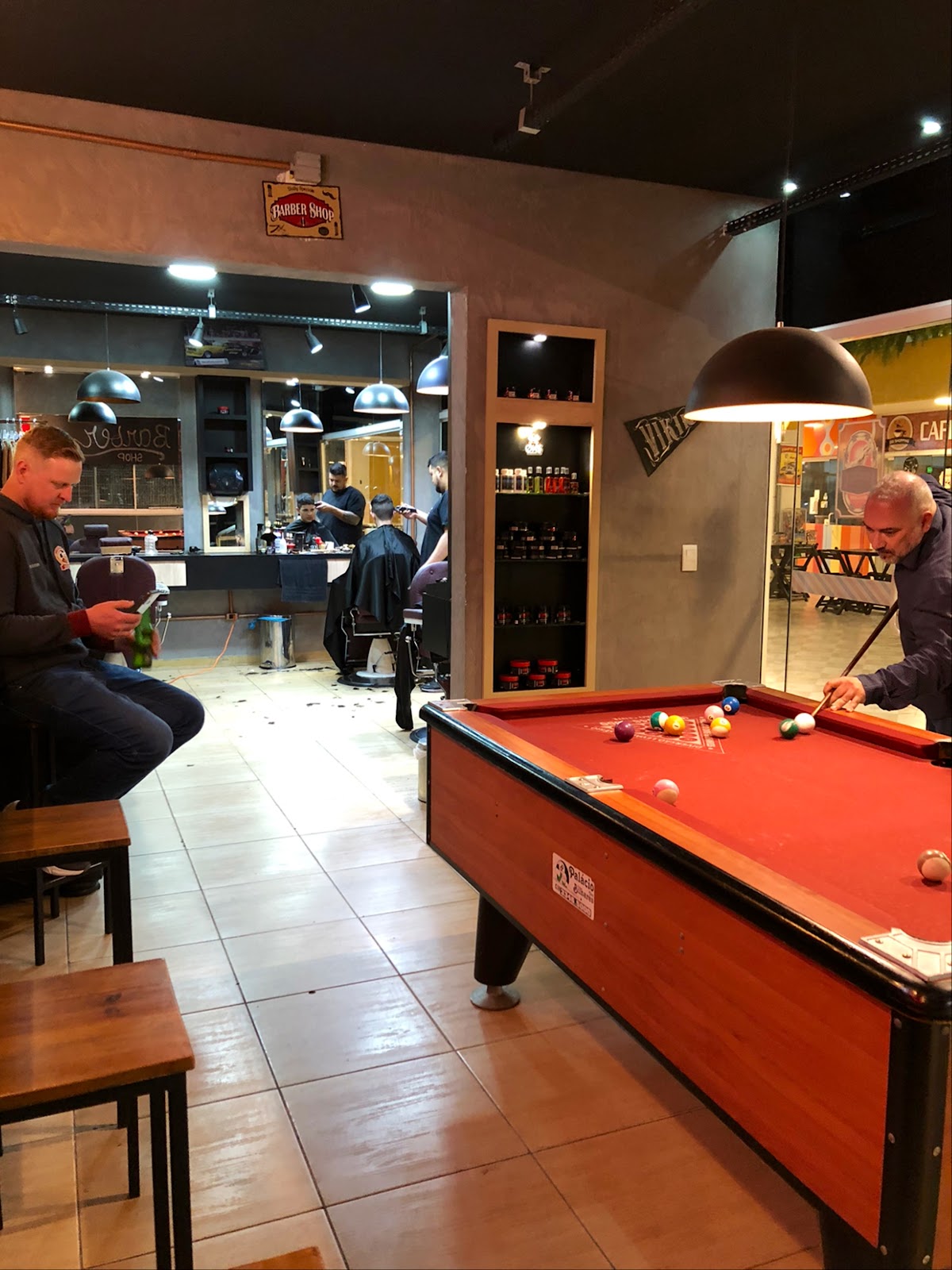 Barbeiros cortando cabelo com mesa de sinuca e área de convivência ao fundo, dentro da Øks Barbearia - Pub